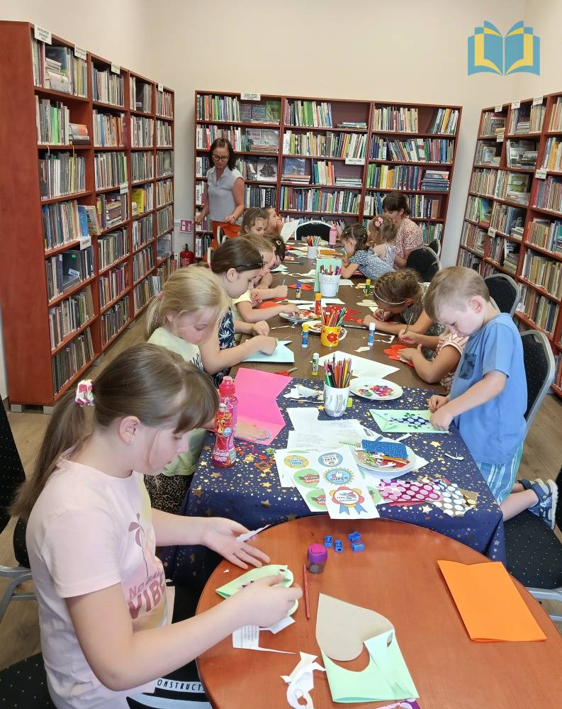 Zdjęcie: Dzieci w bibliotece, siedzą przy długim stole i wykonują laurki na Dzień Taty.